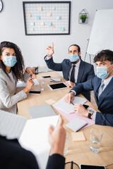 Hispanic office worker with raised hand, sitting near co-workers during business meeting on blurred foreground