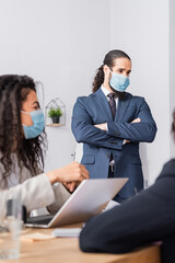 Hispanic businessman in medical mask standing with crossed arms near colleagues on blurred foreground