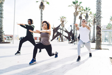 Obraz premium Group of muscular athletes doing squatting exercise during morning workout at urban setting with palm trees, slim female aerobic trainer in bluetooth headphones working with determined male group