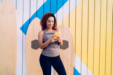 Smiling dark skinned caucasian female in active wear using mobile phone sending text messages and chatting online via app outdoors, cheerful woman blogger share fitness content in social networks