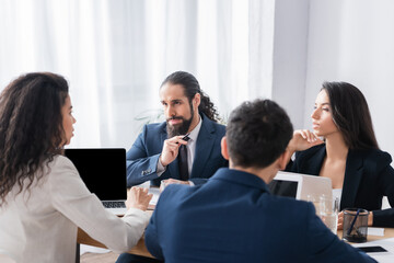 Hispanic businessman looking at colleague near devices on blurred foreground in office