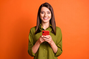 Photo of cheerful positive girl hold cellphone beaming smile look camera wear green shirt isolated orange color background
