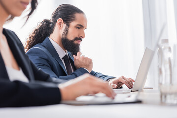 Hispanic businessman using laptop near colleague on blurred foreground in office