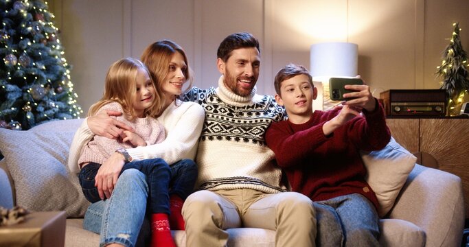 Caucasian cheerful family with children sitting in cozy decorated room with christmas tree and taking selfie photos on smartphone Son taking pictures with parents and sister on New Year's Eve.