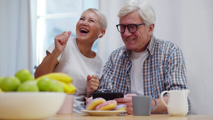 Aged couple listening to music at digital tablet while having delicious breakfast at home kitchen
