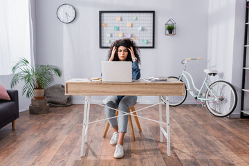 full length of stressed woman adjusting hair while sitting near laptop, smartphone and notebooks on desk