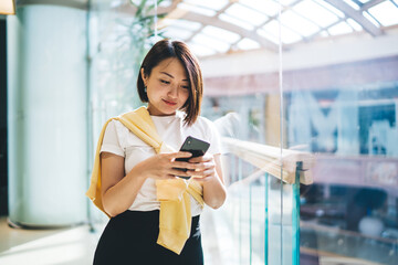 Young Asian female standing in modern building chatting on smartphone smiling gently