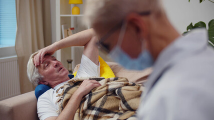 Doctor in mask visiting sick senior woman lying on couch and describing symptoms