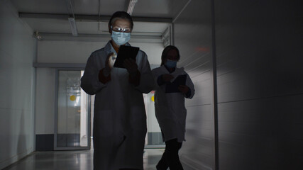 Medical laboratory workers in lab coat and safety mask using digital tablet of clipboard walking in corridor