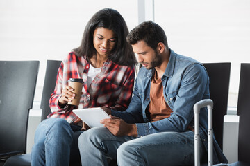 happy african american woman holding disposable cup and looking at digital tablet near bearded man in airport