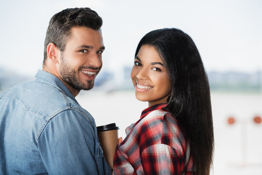 happy multicultural couple smiling near coffee to go in airport
