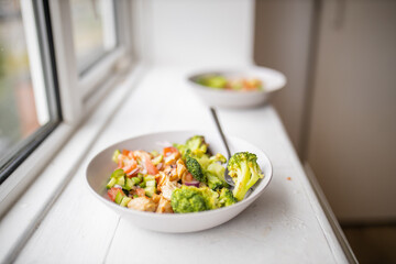 Broccoli and chicken salad on white bowl next to a window