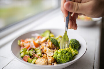 Hand sticking fork on broccoli and chicken salad next to a window