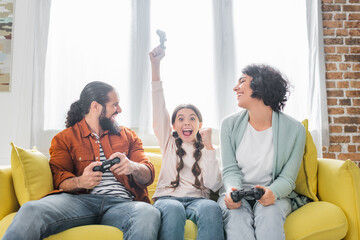 excited hispanic girl showing winner gesture near happy parents holding joysticks