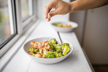 Hand adding a pinch of salt to a broccoli and chicken salad next to a window