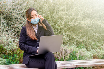 Japanese woman working with her laptop sitting on a park bench, remotely connected Asian...