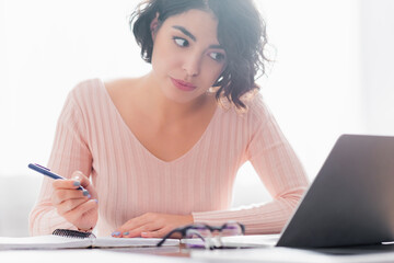 concentrated hispanic woman holding pen while watching webinar on laptop on blurred foreground