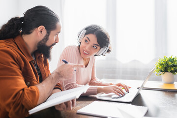 Obraz premium hispanic bearded man pointing with finger near smiling wife in headphones during online learning at home
