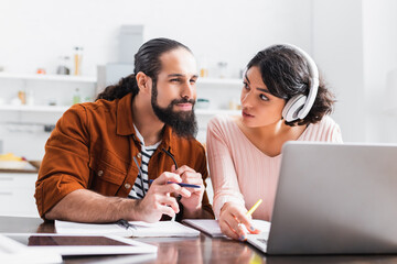 hispanic woman in headphones looking at husband while watching webinar at home