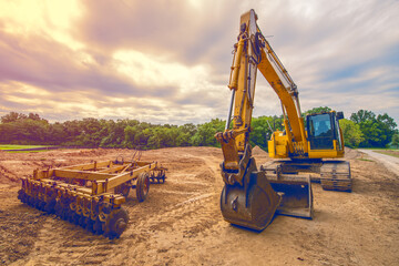 Yellow construction backhoe and disk plow heavy equipment sit in open field after the ground was...