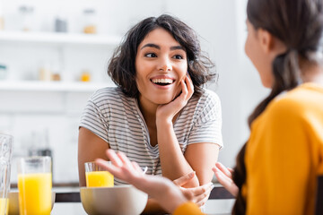 Smiling hispanic woman looking at daughter on blurred foreground during breakfast at home