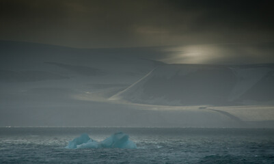 Fototapeta na wymiar A dramatic arctic landscape with mountains lit by a moody sky and in the foreground an iceberg drifts.Sea Ice.