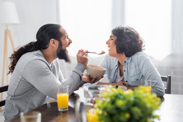 Hispanic man holding spoon of cereals near smiling wife in kitchen
