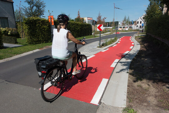 A female cyclist cycles on a dedicated cyle lane which is painted red and runs next to a road.She wears a cycle helmet and has a pannier on her bike