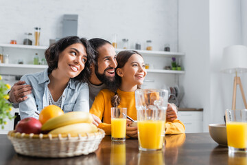 Cheerful hispanic man hugging family near breakfast and orange juice on kitchen table