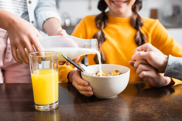 Cropped view of woman pouring milk in cereals near glass of orange juice and family on blurred...