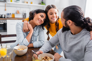 Smiling hispanic girl embracing parents near bowls of cereals on blurred foreground