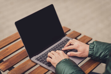 Cropped photo of young guy arms dressed green coat having rest sitting cafeteria typing modern gadget outdoors city street