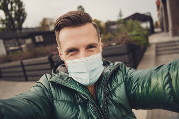 Photo of cute good mood young guy dressed green coat protection mask making selfie outdoors city...
