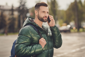 Photo of cheerful cute young guy dressed green coat rucksack walking talking modern gadget outdoors city street