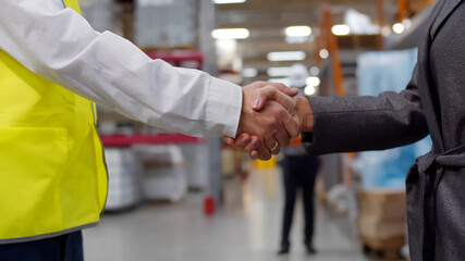 Closeup of businesswoman and industrial worker shaking hands on factory warehouse background