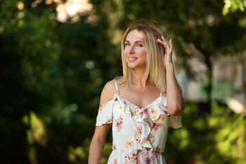 Fototapeta premium Attractive young blonde woman in a light summer dress with flowers, woman enjoying her time outdoors in a summer green park with the sunset in the background