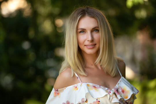 Attractive young blonde woman in a light summer dress with flowers, woman enjoying her time outdoors in a summer green park with the sunset in the background