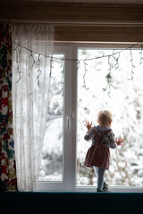 Toddler girl standing on the window decorated for Christmas and watching first snow