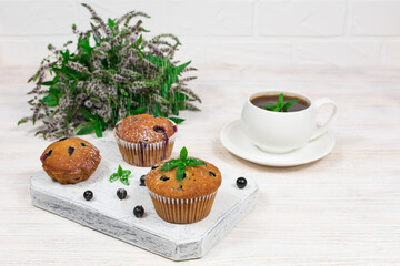 Cupcakes with black currants on a white wooden cutting board against the background of a cup and mint flowers.
