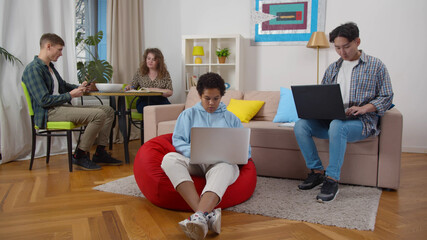 Multiracial group of young friends working on laptop or studying with books in dorm living room