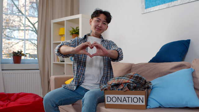 Young Korean Man Sitting On Couch Near Donation Box With Clothes And Gesturing Heart With Hands