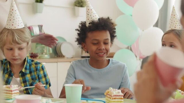 Medium close-up of African boy sitting at table with school friends wearing party hats. Caucasian mother bringing birthday present to son, he looking excited and happy ripping off wrapping paper
