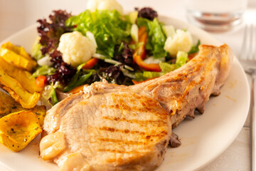 Fried pork entrecote on the bone with stewed pumpkin pieces and salad in a plate close up, green salad with cauliflower and napa cabbage, bell pepper and spices