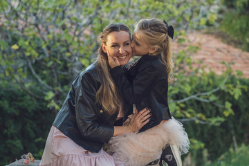 Mom and daughter take a walk in the park in autumn, both wearing pink dresses and black leather jackets, blondes with long hair. Little daughter hugs mom and whispers something in her ear - Powered by Adobe
