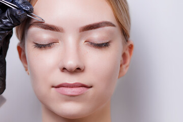 Beautiful young girl tweezing her brows in a beauty salon. Beauty Concept. Cosmetologist plucks client eyebrows by thread. Eyebrow correction, threading. Permanent Makeup. Microblading brow. Close up.