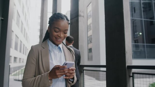 Cheerful young nice-looking black business woman in formal suit using cellphone online communication walking the street city center. Modern lifestyle. Successful youth.