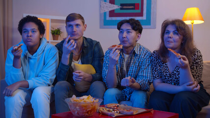 Multiethnic young friends eating pizza and chips watching tv in modern living room