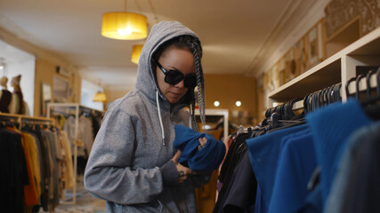 Side view of young woman in sunglasses and hood stealing clothes from store