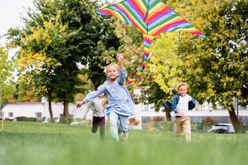 Smiling girl holding flying kite while playing with boys on blurred background in park