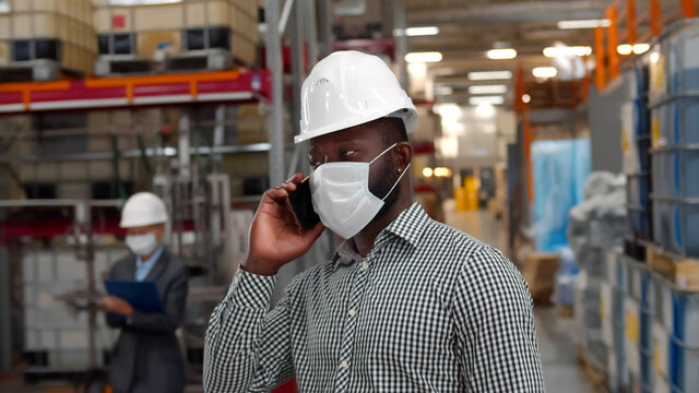 African businessman wearing protective face mask and taking on phone walking in factory warehouse.